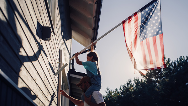 Dad holding his young daughter on his shoulders putting the USA Flag on the wall of their house.
