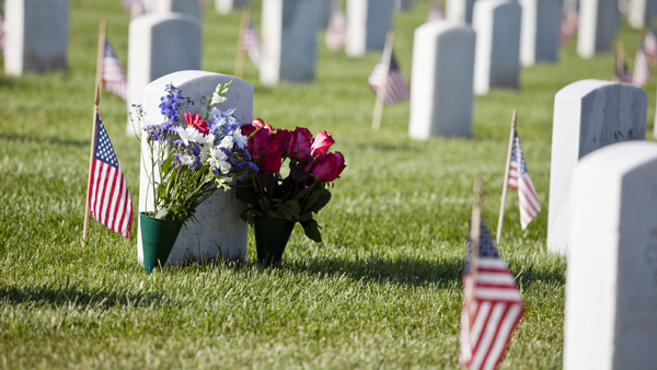Rows of white grave markers on green grass with small U.S. flags planted near each and flowers planted at grave marker in foreground.