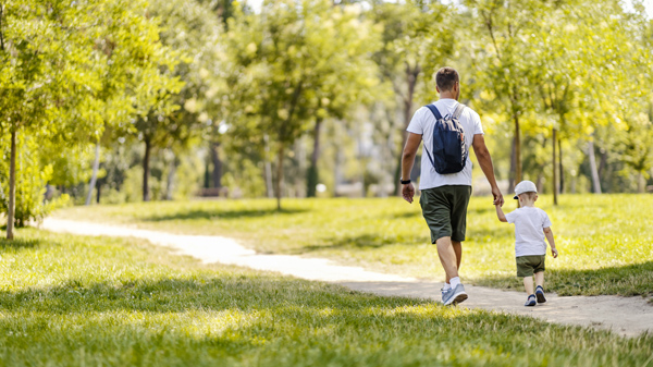 Man wearing backpack holding hand with small son and walking along path in park
