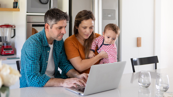 Man types on laptop at kitchen counter while woman watches and holds a baby