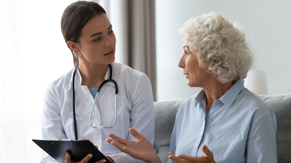 Older female patient in chair speaking to a female doctor holding a chart.