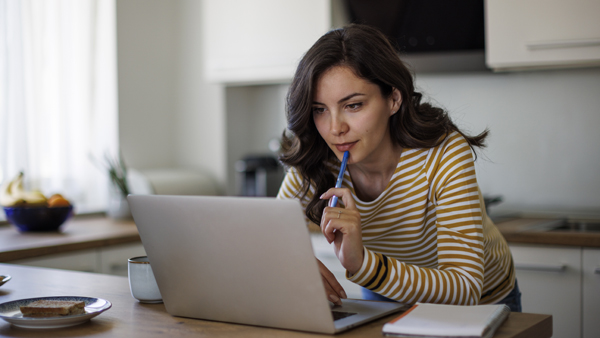 Women looking at laptop computer on kitchen counter while holding a pen and taking notes.