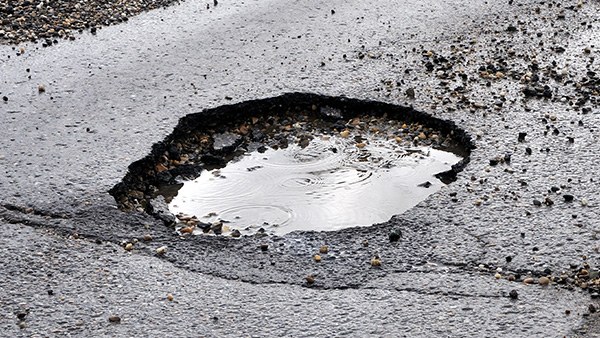 Pothole in asphalt filled with water and surrounded by wet road and gravel.