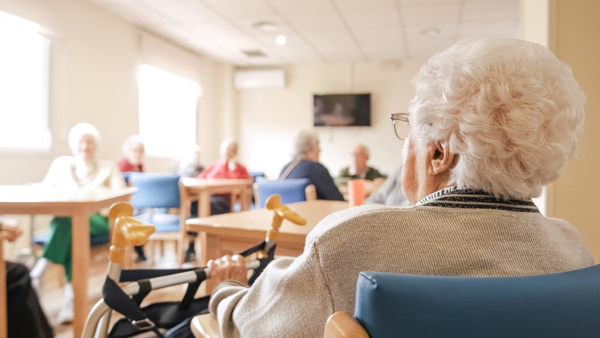 Older woman seated while holding a walker looking at sunny room of senior citizens sitting at tables.