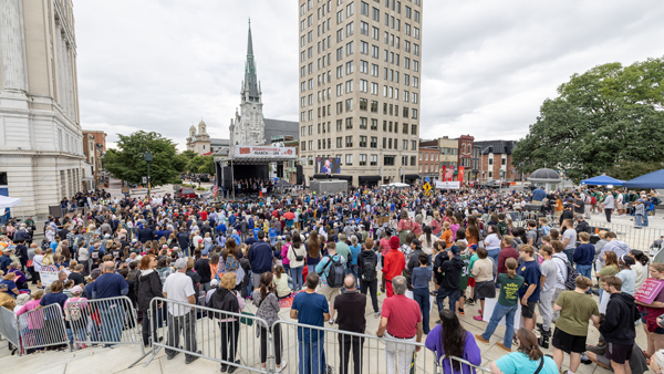 Large crowd on plaza with crowd control gates in the foreground and stage and speakers in background.