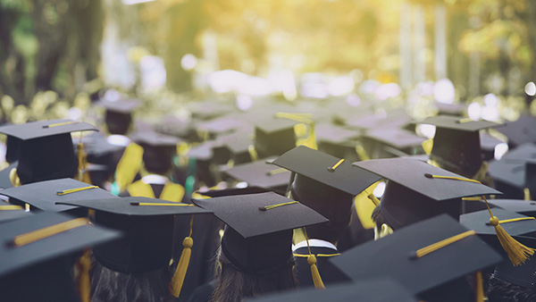Top view of a group of graduates and their caps and tassels at an outdoor graduation ceremony with stage and trees in background.