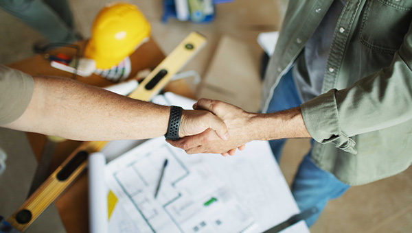 Two men shaking hands over desk covered with blueprints, a level and a hard hat.