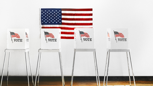 Four voting booths, each with the word “Vote” on it, with U.S. flag on the rear wall.