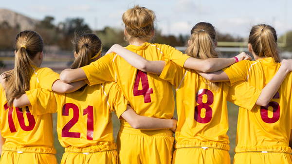 Girl soccer players with arms over each other’s shoulders looking away.